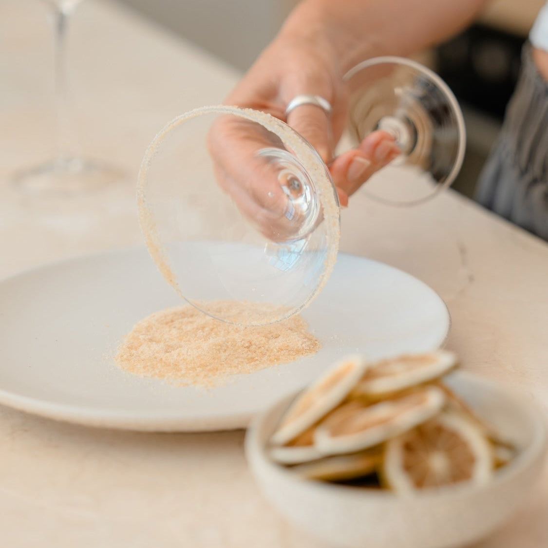 A hand holds cocktail glass over a bed of margarita rim salt on a white plate, with dried lime slices in a small bowl on a light marble countertop. Dipping the rim into the cocktail rim blend.