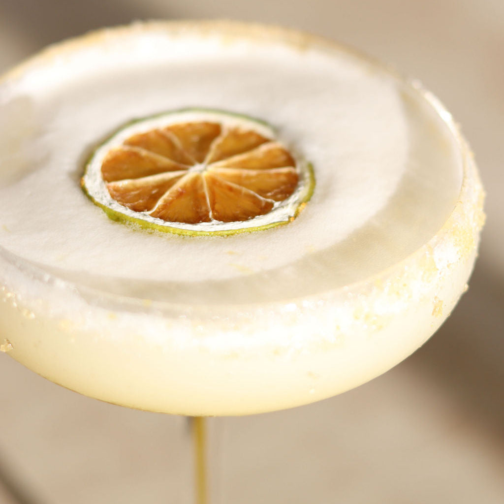 A close-up of a margarita-style cocktail in a salt-rimmed glass with a dried lime slice garnish on top, set against a softly blurred beige background.