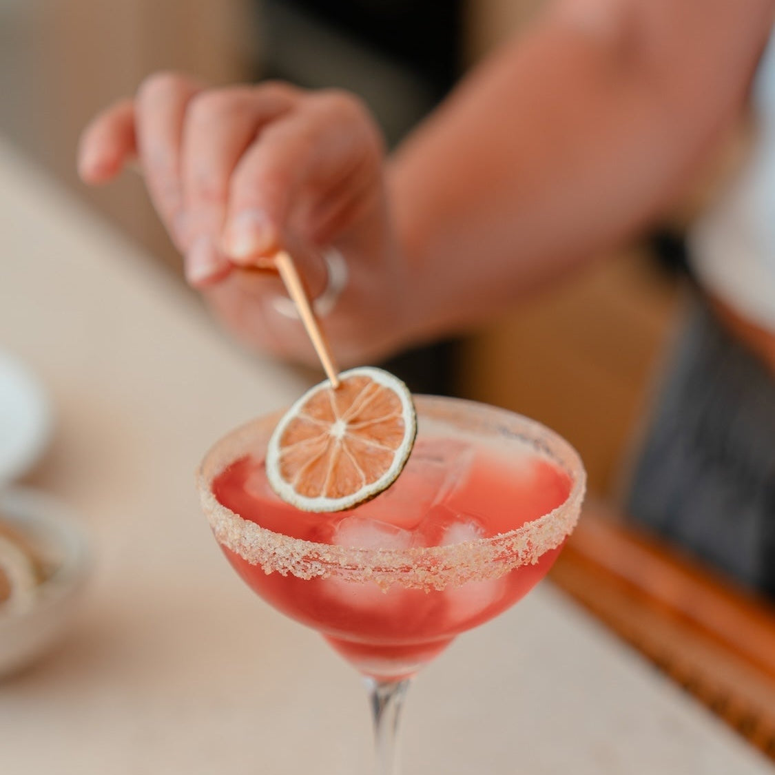 A cranberry margarita cocktail in a coupe glass with a salt/sugar-rimmed edge and a dried lime garnish being placed with garnish tweezers, set on a light stone countertop with a person's hand adjusting the garnish in the background.