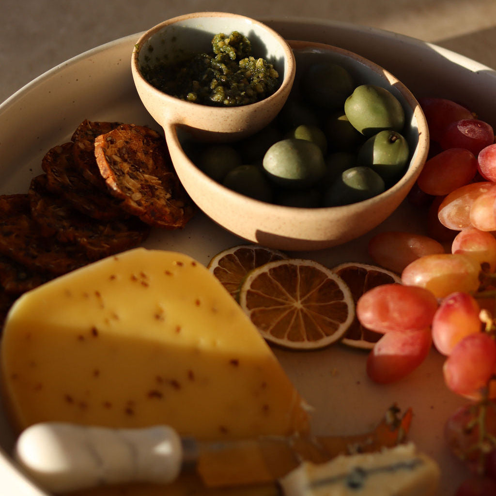 A styled charcuterie board featuring a beige ceramic double-bowl centerpiece with green olives and a small bowl of green spread, alongside a wedge of yellow cheese, red grapes, dried lime slices, and crackers on a matching tray.