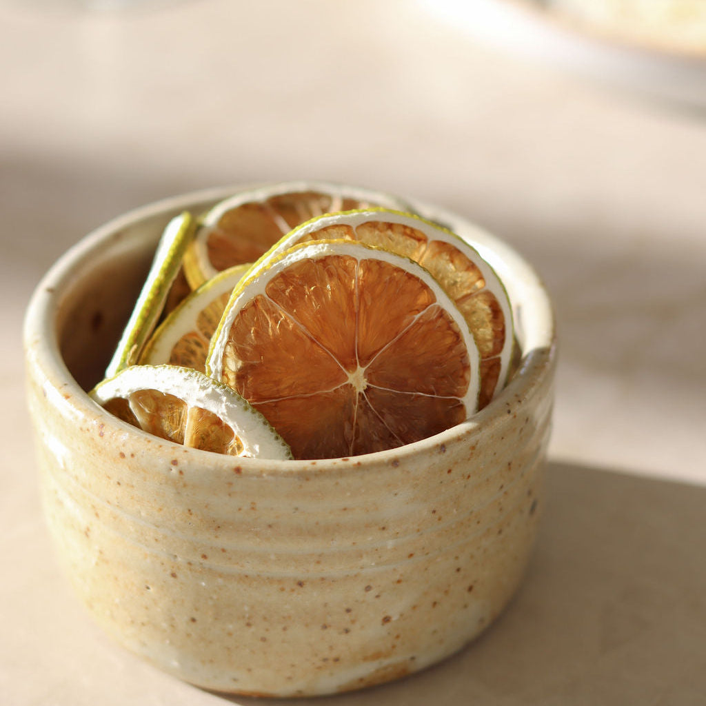 A small speckled ceramic bowl filled with dried lime slices, ready to garnish cocktails, photographed on a warm beige surface with soft sunlight and a blurred background.
