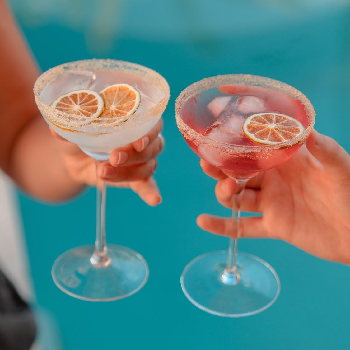 Two hands holding up margarita cocktails in coupe glasses, one with a white drink and two dried lime slices as garnish, and one with a pink drink with dried lime garnish and ice, set against a bright turquoise pool background.  