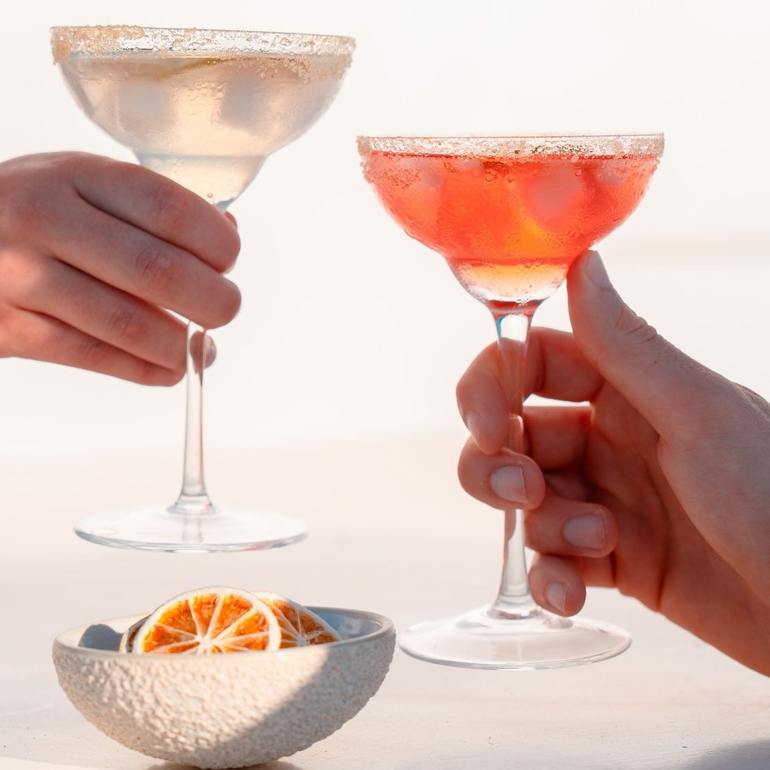 Two hands holding margarita cocktail glasses at a beach, with one glass containing a clear pale drink and the other a pink-orange drink, against a bright white background. A bowl of dried lime slices in the foreground. 