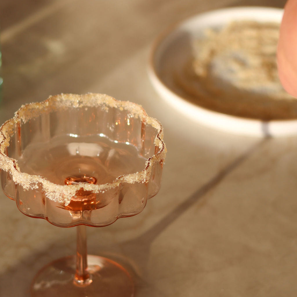 A close-up of a pink cocktail glass coupe-style cocktail glass with a scalloped rim coated in lime rim blend salt and sugar, set on a beige surface with a shallow depth of field and a blurred plate in the background.