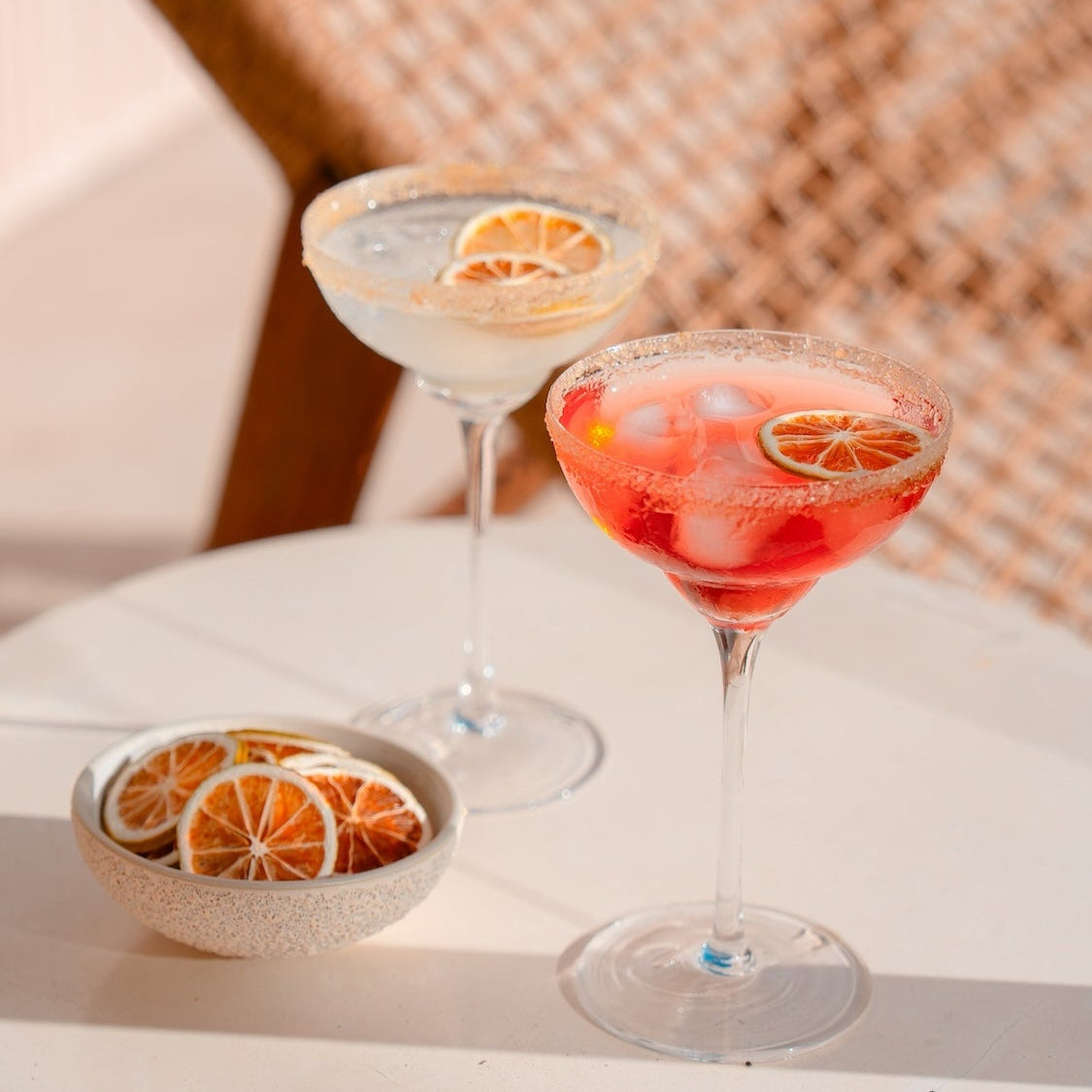 Two margarita cocktails in coupe glasses on a white table, one clear and one pinkish-red, each garnished with a dried lime slice, with a small bowl of dried lime slices in the foreground and a woven chair blurred in the background.