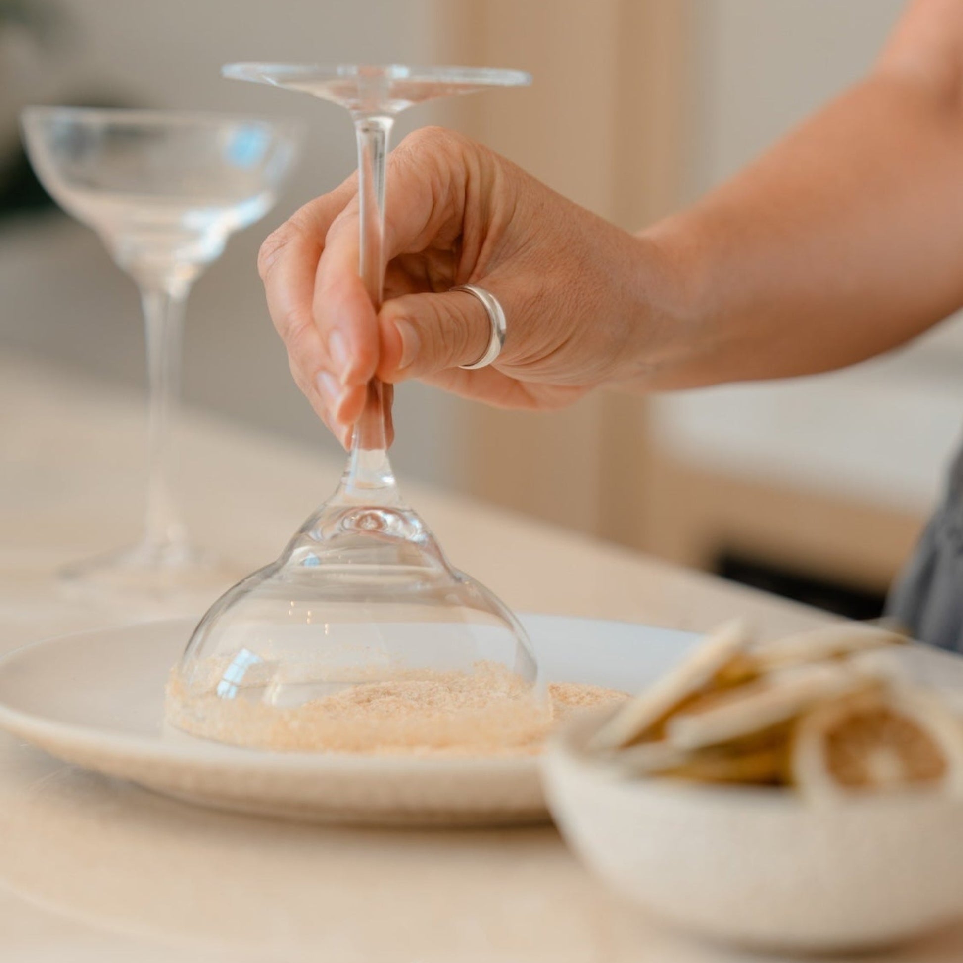 A close-up lifestyle shot of a person's hand dipping a clear glass cocktail glass into cocktail rim salt blend on a white plate, with a small bowl of dried lime in the foreground on a light beige countertop.