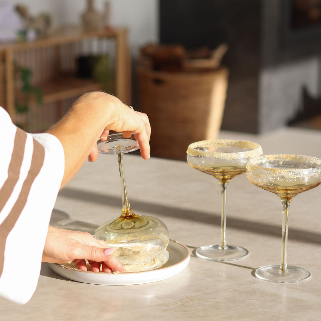 A person's hands are shown placing a margarita glass into the lime rim blend on a small white plate, with two rimmed cocktail coupe glasses standing nearby on a light stone countertop in a kitchen setting.