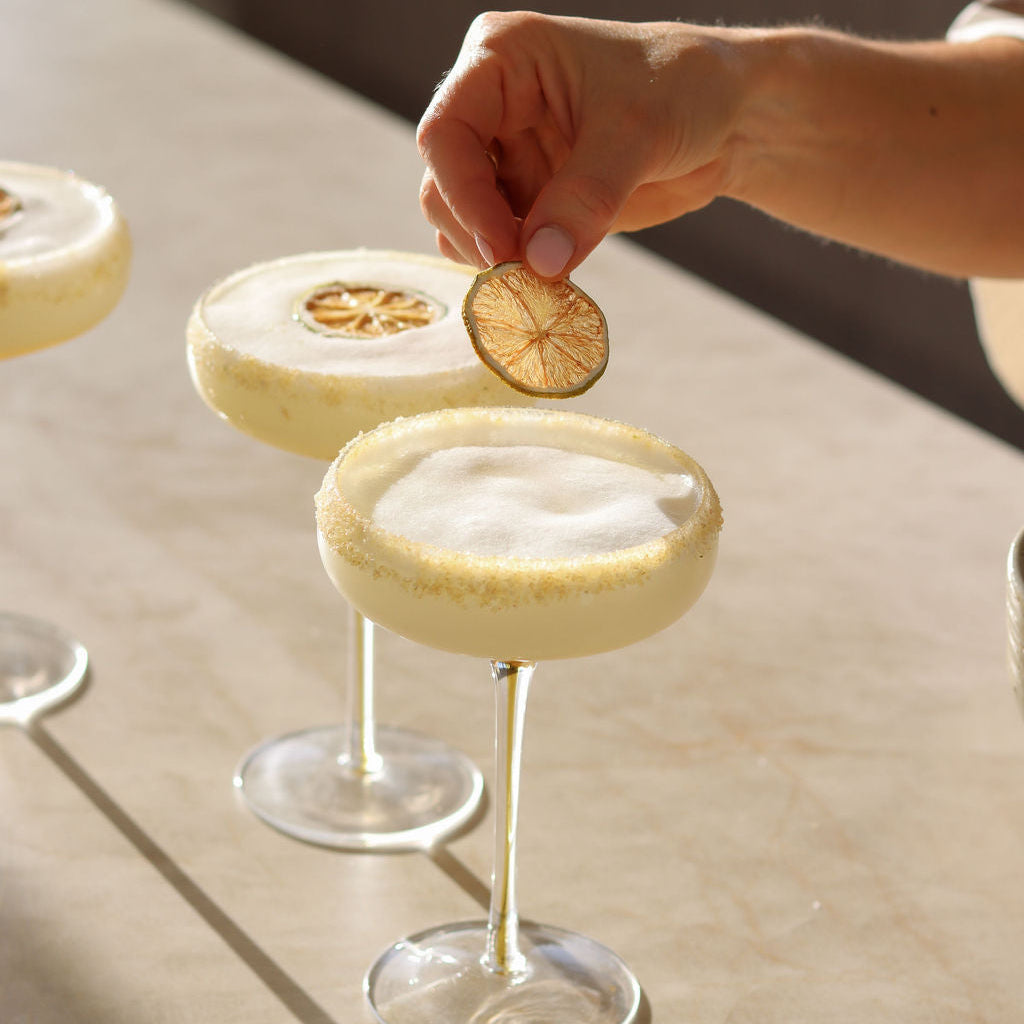A hand garnishes a creamy coconut margarita cocktail in a coupe glass with a dried lime slice, with two more cocktails blurred in the background on a sunlit table.